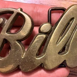 Puede incluir: Una hebilla de cinturón de latón con el nombre Bill en una fuente cursiva en relieve. El metal tiene un acabado pulido y ligeramente desgastado con una púa de metal marrón oscuro en la parte posterior para asegurar la hebilla a la correa.