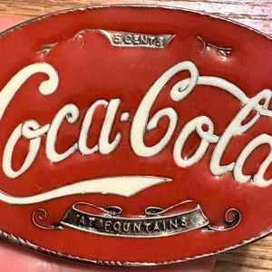 Puede incluir: Una hebilla de cinturón ovalada de metal rojo y blanco con el logotipo de Coca-Cola en letras blancas. La banda superior dice 5 CENTS y la banda inferior dice AT FOUNTAINS. El diseño tiene un acabado brillante con un borde metálico plateado.