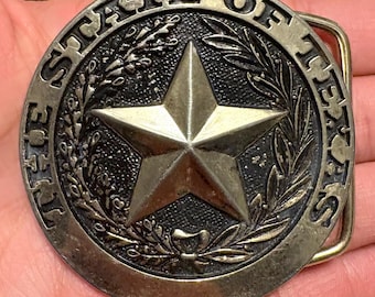 May include: A round, antique-style belt buckle with the words "THE STATE OF TEXAS" encircling a central star. The star is surrounded by a wreath design. The buckle is made of a bronze-colored metal.