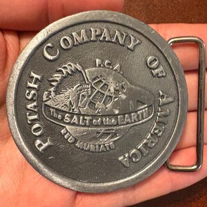 Op de afbeelding: Een cirkelvormige, zilverkleurige metalen riemgesp. De gesp heeft de tekst "POTASH COMPANY OF AMERICA" rond de rand. Het middenontwerp bevat een wereldbol en de woorden "The SALT of the EARTH" en "RED MURIATE".
