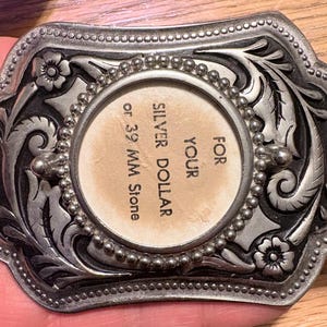 Puede incluir: Hebilla de cinturón de metal plateado con un diseño floral y de volutas ornamentado. El círculo central está diseñado para contener un dólar de plata o una piedra de 39 mm. La hebilla tiene un borde con cuentas y se sostiene en una mano.