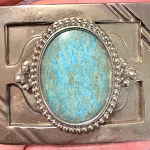 Può includere: Fibbia per cintura rettangolare color argento con una pietra turchese ovale al centro. La pietra è circondata da una cornice argentata con perline. La fibbia presenta dettagli incisi e un classico design western.
