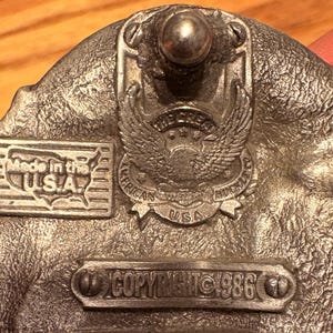 May include: A silver-colored belt buckle with an eagle emblem and the text "American Buckle USA." The buckle also features the text "Made in the USA" and "Copyright 1986."