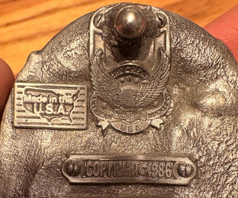 May include: A silver-colored belt buckle with an eagle emblem and the text "American Buckle USA." The buckle also features the text "Made in the USA" and "Copyright 1986."