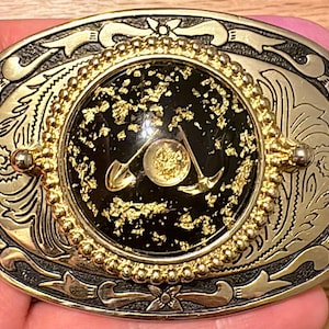Pode incluir: Fivela de cinto ovalada dourada e prateada com um centro preto. O centro apresenta flocos de ouro e um design de pá e picareta dourados. A fivela tem uma borda ornamentada e gravada com detalhes florais e de rolagem.