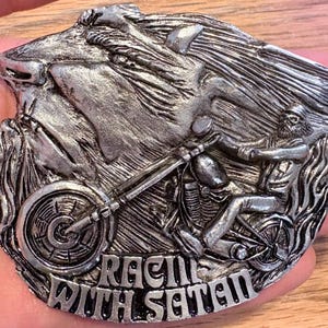 May include: Silver-toned metal belt buckle featuring a biker riding a motorcycle, with a wolf's head design. The text "RACING WITH SATAN" is displayed at the bottom. The buckle has intricate details and a vintage aesthetic.