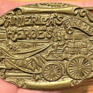 Puede incluir: Hebilla de cinturón de latón vintage con las palabras "America's Heroes Fire Dept" en relieve. La hebilla presenta un relieve detallado de un carro de bomberos tirado por caballos con bomberos. La hebilla es de forma ovalada.