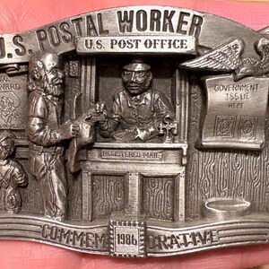 Puede incluir: Hebilla de cinturón de metal plateado con temática de trabajador postal. La hebilla tiene imágenes en relieve detalladas de trabajadores postales, un entorno de oficina de correos y texto que dice "U.S. Postal Worker" y "Commemorative 1986". Incluye un águila y un pergamino "Government Issue".