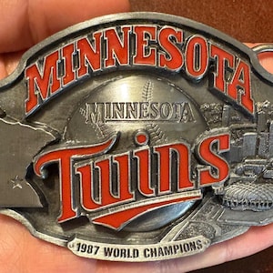 May include: A silver-colored belt buckle featuring the text "MINNESOTA" in red, arched across the top. Below, the word "Twins" is in red, with a baseball graphic in the center. The buckle also includes the text "1987 WORLD CHAMPIONS".