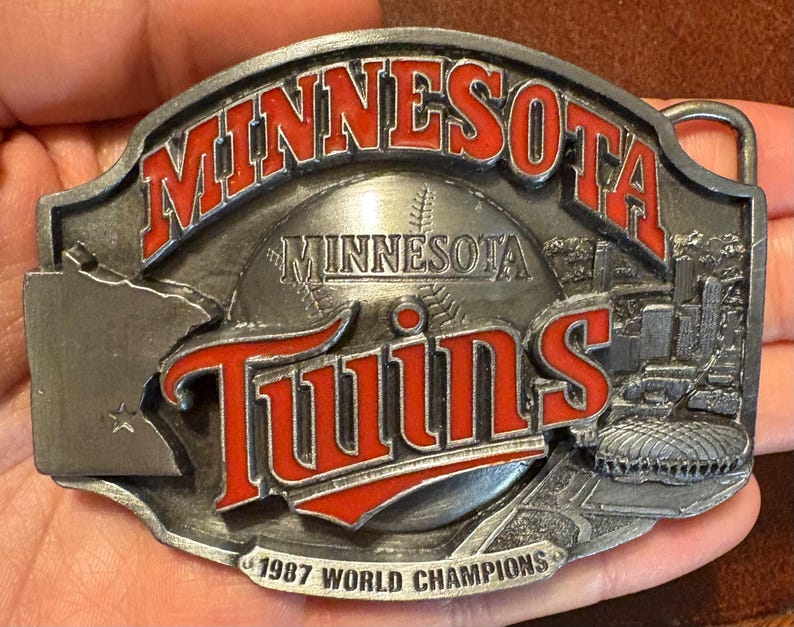 May include: A silver-colored belt buckle featuring the text "MINNESOTA" in red, arched across the top. Below, the word "Twins" is in red, with a baseball graphic in the center. The buckle also includes the text "1987 WORLD CHAMPIONS".