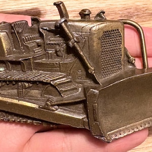 Könnte beinhalten: Detaillierte, bronzefarbene Gürtelschnalle in Form eines Vintage-Bulldozers. Die Schnalle weist komplizierte Details der Ketten, des Schildes und der Motorkomponenten des Fahrzeugs auf. Die Schnalle wird in einer Hand gehalten.