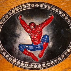 Peut inclure: Une boucle de ceinture ovale en métal présentant une figurine en relief émaillée d'un super-héros rouge et bleu dans une pose accroupie dynamique. La figure centrale est sur fond noir lisse, entourée d'une bordure de strass ronds transparents.