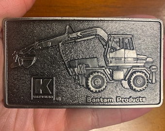 Puede incluir: Hebilla de cinturón rectangular plateada con una imagen en relieve de una excavadora Bantam Products. La hebilla tiene el logotipo de Kobring y las palabras "Bantam Products" son visibles.