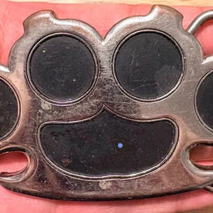 May include: A silver-colored belt buckle with a brass knuckle design. The buckle has five circular black insets and a rectangular black inset at the bottom. The buckle is held in a hand.