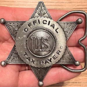 May include: A silver-colored metal belt buckle in the shape of a six-pointed star. The center circle reads "OFFICIAL US TAX PAYER". The buckle has a decorative design and a curved metal loop for the belt.