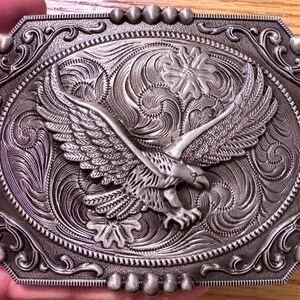 May include: A rectangular silver-toned metal belt buckle featuring a raised eagle with wings spread wide in the center. The background is decorated with intricate engraved scrollwork and floral patterns, framed by a border of small silver spheres.