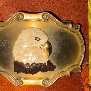 May include: A vintage brass belt buckle featuring a detailed eagle head. The eagle is cream-colored with a black neck and a gold beak, set against a gold-toned oval frame. The buckle measures approximately 3 inches in length.