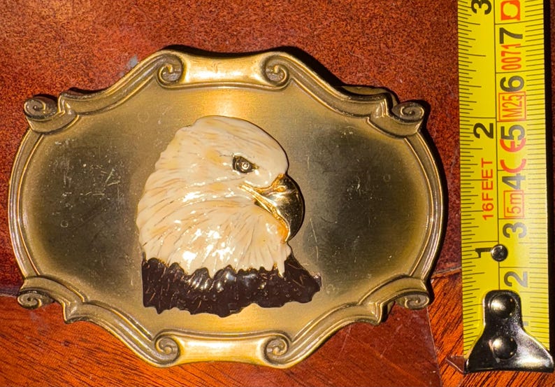 May include: A vintage brass belt buckle featuring a detailed eagle head. The eagle is cream-colored with a black neck and a gold beak, set against a gold-toned oval frame. The buckle measures approximately 3 inches in length.