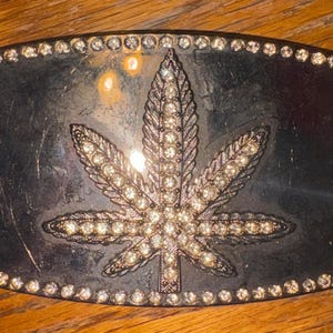 May include: A rectangular belt buckle with a dark metallic finish, featuring a central cannabis leaf motif encrusted with clear rhinestones. The perimeter of the buckle is also lined with a continuous border of small, clear, sparkling rhinestones.