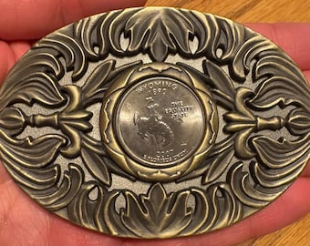 May include: An oval-shaped, antique brass belt buckle with an ornate floral design. The centre features a Wyoming quarter coin. This vintage accessory is ideal for adding a touch of Western style to any outfit.