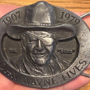Può includere: Fibbia per cintura in metallo color argento con un ritratto di John Wayne con cappello da cowboy e benda sull'occhio. La fibbia è di forma ovale con le scritte "1907" e "1979" in alto e "JOHN WAYNE LIVES" in basso. È visibile anche la parola "DUKE".