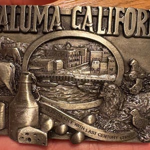 Może przedstawiać: Srebrna klamra do paska z napisem "PETALUMA CALIFORNIA" na górze. Klamra przedstawia szczegółowy relief sceny miejskiej, zwierząt gospodarskich i butelki. Obecny jest również tekst "A MODERN CITY WITH LAST CENTURY CHARM".