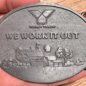 May include: Oval silver-coloured belt buckle with the text "WE WORK IT OUT" and "NODER FEEDS". The buckle features a rural scene with a barn, tree, and moon. The buckle has a metal loop for the belt.