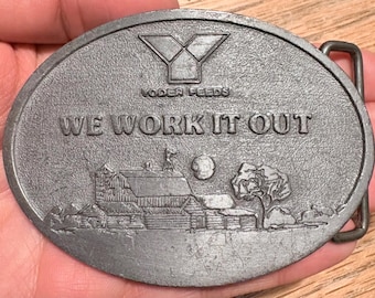 Op de afbeelding: Ovale, zilverkleurige gesp met de tekst "WE WORK IT OUT" en "NODER FEEDS". De gesp toont een landelijke scène met een schuur, boom en maan. De gesp heeft een metalen lus voor de riem.
