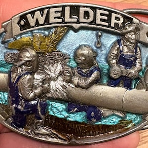 May include: Oval metal belt buckle with a detailed relief scene of welders at work. The word "WELDER" is across the top. The buckle features figures in blue overalls, welding equipment, and a blue and green background.