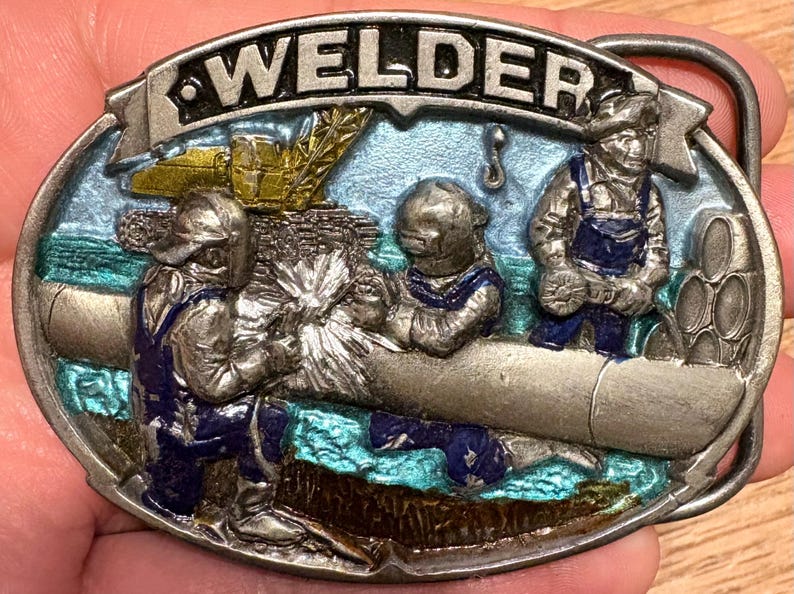 May include: Oval metal belt buckle with a detailed relief scene of welders at work. The word "WELDER" is across the top. The buckle features figures in blue overalls, welding equipment, and a blue and green background.