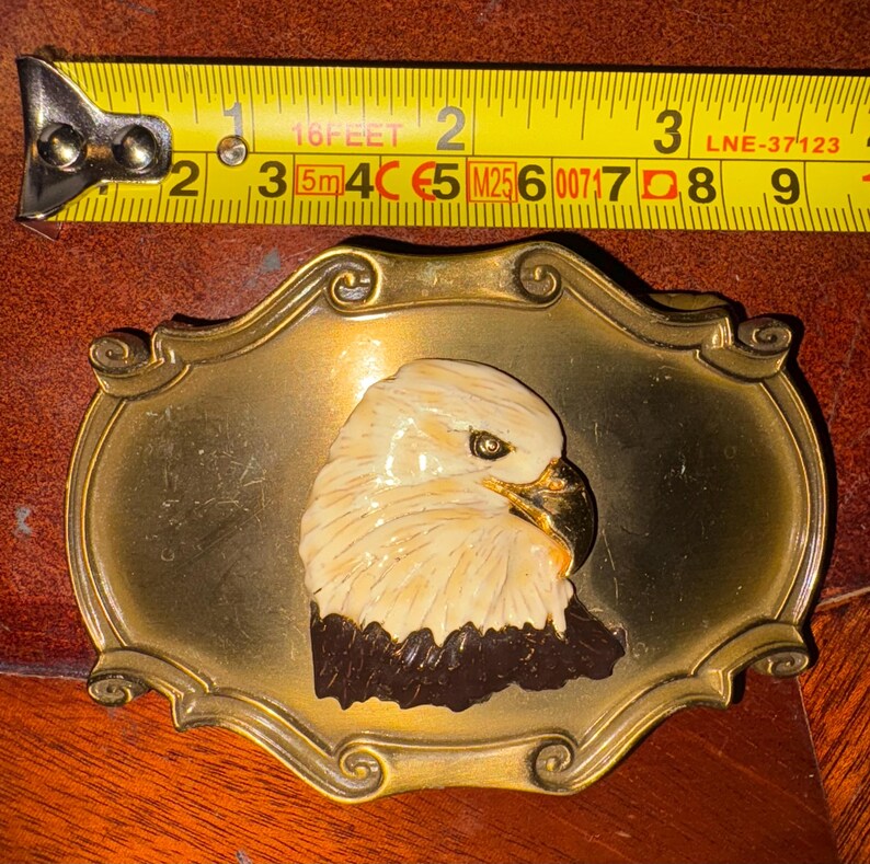 May include: Vintage brass belt buckle featuring a sculpted bald eagle head. The eagle has a cream-colored head and a dark brown neck, set against a gold-toned rectangular frame. The buckle measures approximately 3 inches in length.