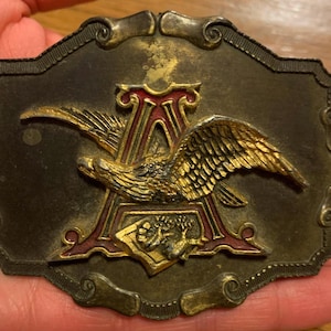 May include: A gold-toned belt buckle with an eagle design. The eagle is perched on top of a red and gold letter 'A'.