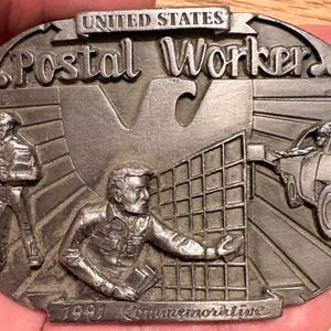 以下が含まれることがあります： UNITED STATES POSTAL WORKERおよび1991 Commemorativeの浮き彫り文字が施された、銀色の楕円形金属製ベルトバックル。歩く郵便局員、格子状の棚で郵便物を仕分ける郵便局員、そして郵便配達車両がデザインされています。