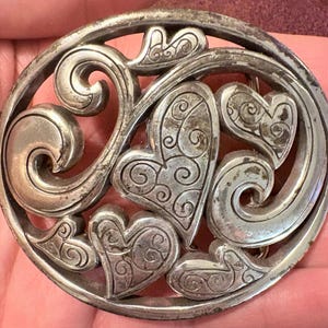 Pode incluir: Um broche circular prateado com um design vazado. O broche apresenta múltiplas formas de coração e padrões em espiral em forma de volutas dentro de uma moldura circular. O metal tem uma aparência ligeiramente envelhecida.