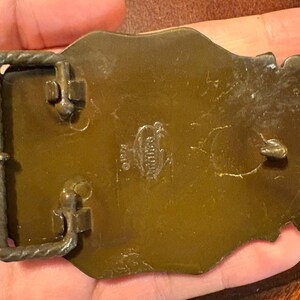 May include: A vintage brass belt buckle with a rectangular frame and a decorative design. The buckle has a tarnished, aged appearance, with visible wear and tear. The back of the buckle has stamped text.