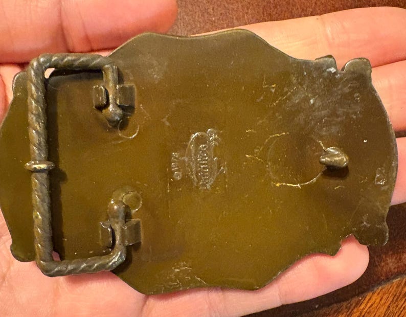May include: A vintage brass belt buckle with a rectangular frame and a decorative design. The buckle has a tarnished, aged appearance, with visible wear and tear. The back of the buckle has stamped text.
