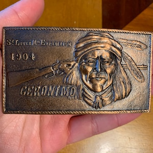 May include: A rectangular bronze-colored belt buckle featuring a detailed relief of Geronimo, a Native American leader, with a rifle. The buckle is inscribed with "St. Louis Mo. Exposition 1904" and the name "GERONIMO".