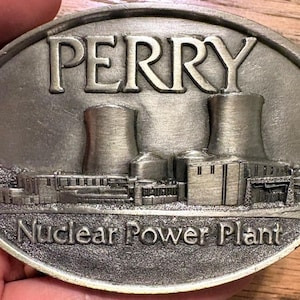Puede incluir: Una hebilla de cinturón ovalada de metal plateado con un relieve de una central nuclear. La palabra PERRY está grabada en letras mayúsculas grandes en la parte superior y el texto Nuclear Power Plant está grabado en la parte inferior.