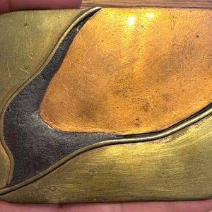 May include: A rectangular metal belt buckle with rounded corners, featuring a multi-toned surface. The design consists of three distinct sections in brushed gold, polished copper, and a dark, textured charcoal gray, separated by raised metal lines.
