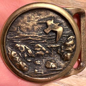 May include: Vintage brass belt buckle with a detailed relief scene of a bird in flight over water and rocks. The buckle has a circular design with a rectangular bar for the belt. The metal has a tarnished, antique finish.
