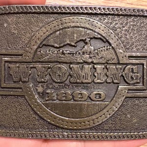 May include: A rectangular, antique-style metal belt buckle with the word "WYOMING" in raised letters. The buckle features a circular design with a landscape scene and the year "1890". The buckle has a textured, aged appearance.