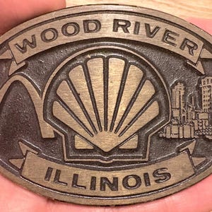 May include: Oval-shaped, bronze-colored belt buckle featuring the words "WOOD RIVER" and "ILLINOIS". The design includes an arch, a stylized shell, and a cityscape. The buckle is held in a hand.