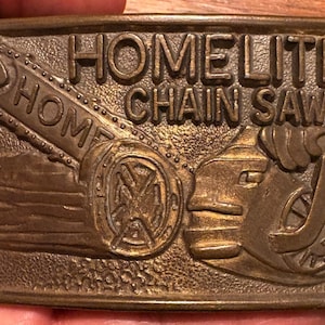 Op de afbeelding: Een rechthoekige messingkleurige metalen riemgesp met een reliëfontwerp van een Homelite-kettingzaag. De tekst HOMELITE CHAIN SAWS staat bovenaan in reliëf. Het ontwerp bevat een gedetailleerde weergave van het zaagblad en de motorbehuizing.
