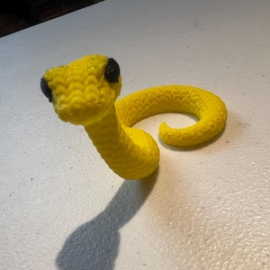 May include: A yellow crocheted snake toy with black eyes. The snake is curled up in a coil.