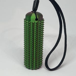 May include: A green and purple cylindrical pendant with a textured, spiky surface. The pendant is attached to a black cord. The design resembles a cactus. The background is plain white.