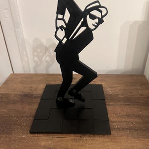 May include: Black silhouette of a person in a suit and hat, standing on a black square base. The figure is posed with one arm raised and the other bent at the elbow.