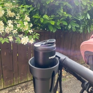 May include: Black stroller cup holder with a black insulated water bottle inside. The cup holder has a black strap.