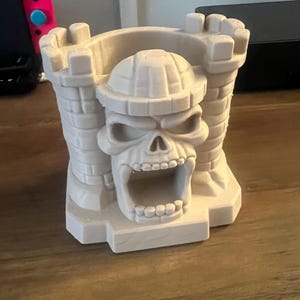 May include: A light-coloured, 3D-printed skull-shaped castle. The castle has a large open mouth, a detailed skull face, and a crenellated top. The design is reminiscent of a fortress or a fantasy-themed structure. The object is sitting on a wooden surface.