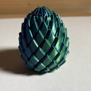 May include: A teal and blue metallic decorative egg. The surface is divided into diamond-shaped segments, giving it a textured look. The object is on a white surface, with a shadow to the left. The egg is approximately 8 cm tall.