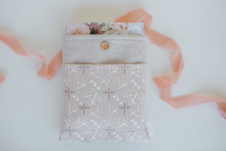 Puede incluir: Una bolsa de tela beige con cierre de bot&oacute;n, con un estampado de corazones blancos, cruces y la palabra "Jes&uacute;s". La bolsa contiene tarjetas con dise&ntilde;os florales. Una cinta melocot&oacute;n est&aacute; drapeada cerca.